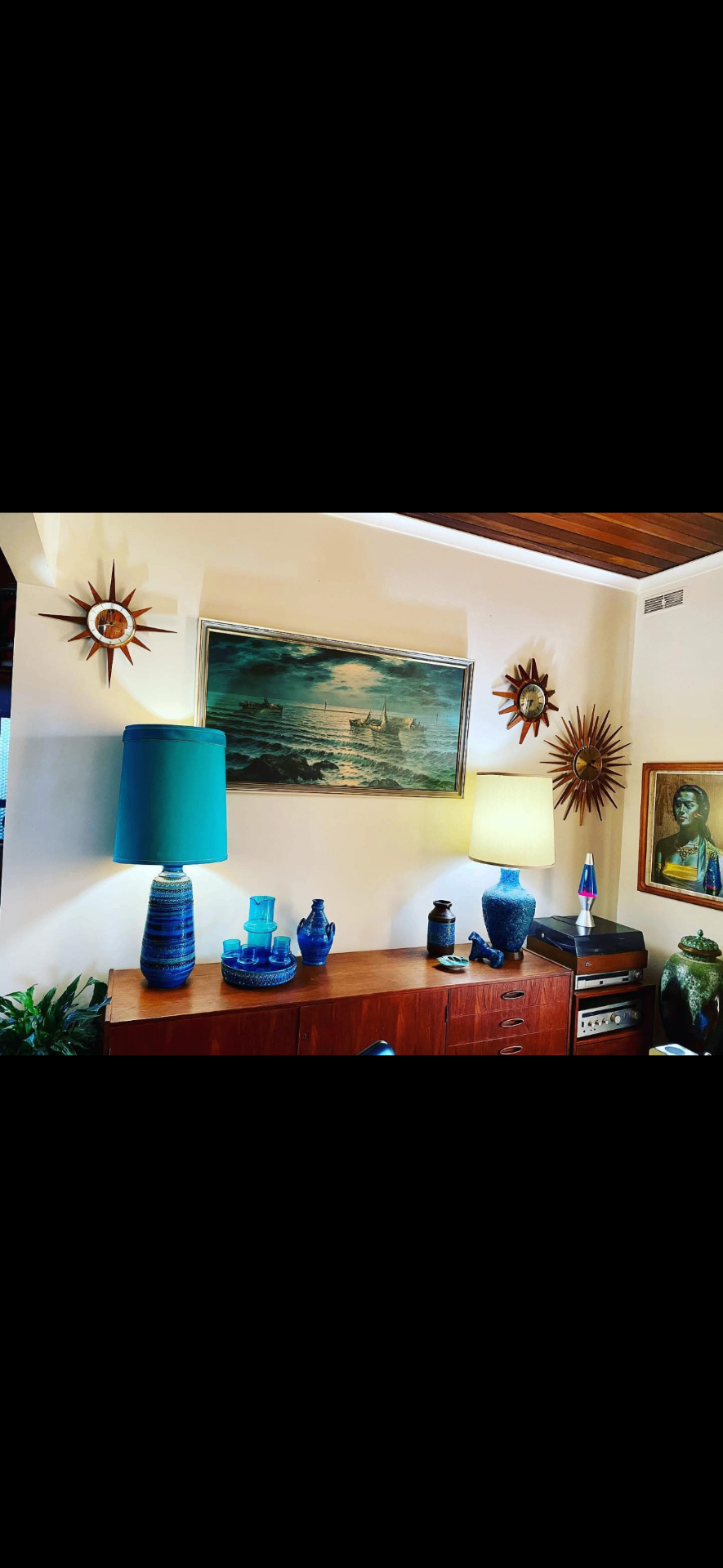 Two teal ceramic lamps glowing on a teak sideboard, with a vintage seascape painting and three starburst clocks on the wall.