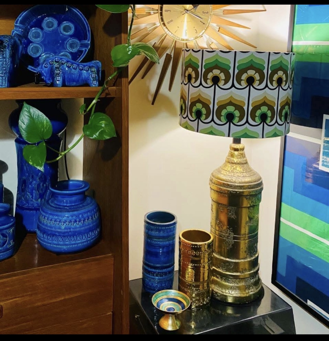 Grouping of Bitossi-style ceramic pieces in gold glaze with a patterned drum-shade lamp and cobalt vases on a teak sideboard.